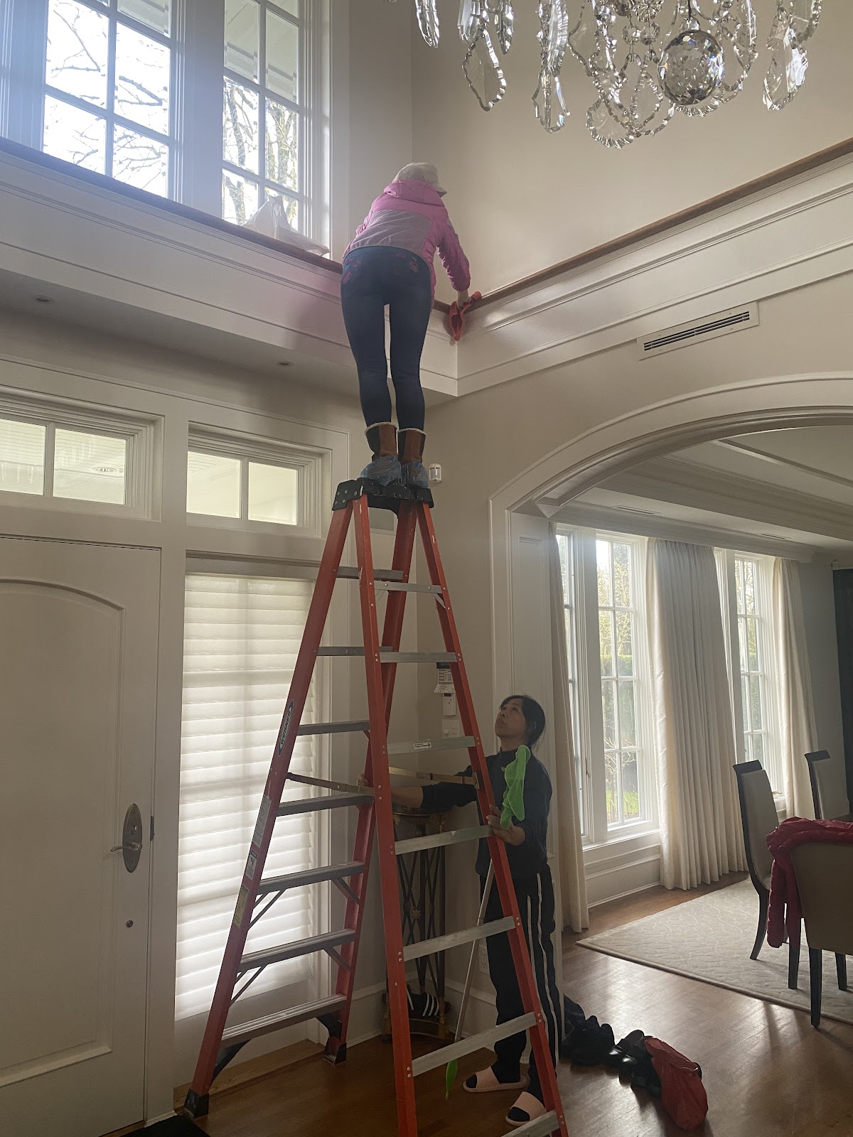 Professional cleaner on tall ladder reaching high areas in an elegant home with chandelier
